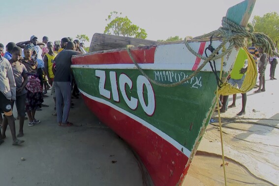 In this video grab provided by Television Mozambique, people stand on Monday, April 8, 2024, next to a boat that local media say capsized on Sunday off Mozambique's northern coast in Nampula province. The makeshift ferry reportedly was overcrowded with residents fleeing a feared cholera outbreak when it capsized, killing at least 98 people including children, media reports say. (Television Mozambique via AP)