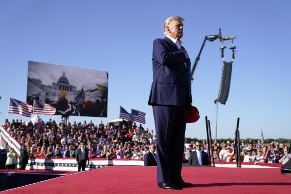 As footage from the Jan. 6, 2021, insurrection at the U.S. Capitol is displayed in the background, former President Donald Trump stands while a song, "Justice for All," is played during a campaign rally at Waco Regional Airport, Saturday, March 25, 2023, in Waco, Texas. The song features a choir of men imprisoned for their role in the Jan. 6, 2021, insurrection singing the national anthem and a recording of Trump reciting the Pledge of Allegiance. (AP Photo/Evan Vucci)