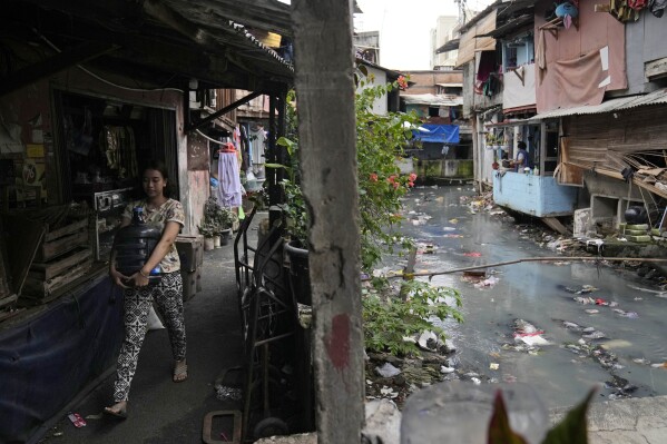 A woman carries a bottle of drinking water she just bought at a nearby store, at a lower-income neighborhood along a polluted canal in Jakarta, Indonesia, Wednesday, March 13, 2024. Access to clean water is uncertain and millions of Indonesians across the country buy water in large refillable containers or single-use packaged plastic bottles. (AP Photo/Dita Alangkara)