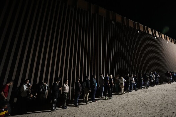 FILE - People line up against a border wall as they wait to apply for asylum after crossing the border from Mexico. Tuesday, July 11, 2023, near Yuma, Ariz. As the number of migrants coming to the U.S.'s southern border is climbing, the Biden administration aims to admit more refugees from Latin America and the Caribbean over the next year. The White House Friday, Sept. 29, released the targets for how many refugees it aims to admit over the next fiscal year starting October 1 and from what regions of the world. (AP Photo/Gregory Bull, File)