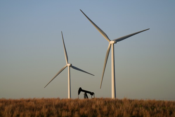 An oil pumping unit works in the foreground while wind turbines at the Buckeye Wind Energy wind farm rise in the distance Sept. 30, 2024, near Hays, Kan. (AP Photo/Charlie Riedel, File)