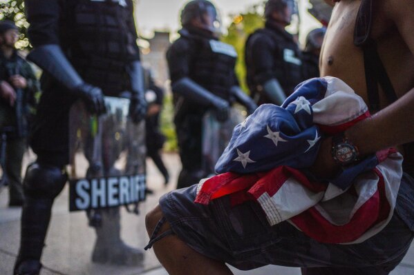 A protester kneels while holding a U.S. flag in front of law enforcement officers in Grand Rapids, Mich., Wednesday, June 3, 2020. (Anntaninna Biondo/The Grand Rapids Press via AP)