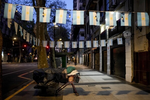 A homeless person sleeps on a bed made of a shopping cart and bench on a sidewalk decorated with Argentine flags in Buenos Aires, Argentina, early Wednesday, Sept. 27, 2023. (AP Photo/Natacha Pisarenko)