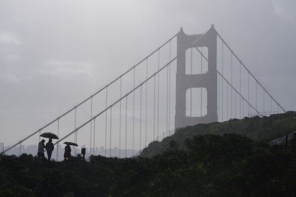 People walk in the rain as a storm moves through with the Golden Gate Bridge in the background near Sausalito, Calif., Monday, Feb. 5, 2024. In Northern California, the storm inundated streets and brought down trees and electrical lines Sunday throughout the San Francisco Bay Area, where winds topped 60 mph (96 kph) in some areas. (AP Photo/Eric Risberg)