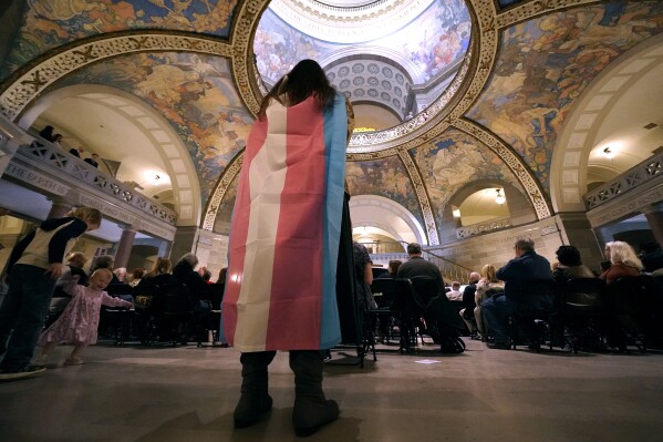 FILE - Glenda Starke wears a transgender flag as a counter protest during a rally in favor of a ban on gender-affirming health care legislation, March 20, 2023, at the Missouri Statehouse in Jefferson City, Mo. A Missouri judge said Friday, Aug. 25, that a law banning gender-affirming treatments for minors can take effect. St. Louis Circuit Judge Steven Ohmer ruled that the law will kick in Monday, Aug. 28, as previously scheduled. (AP Photo/Charlie Riedel, File)