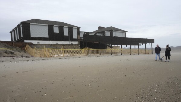 Passers-by walk a dog past a damaged structure on Dunes Club Beach, Thursday, Jan. 25, 2024, in Narragansett, R.I. Experts say erosion and receding shorelines are becoming more common due to ocean rise and climate change. (AP Photo/Steven Senne)