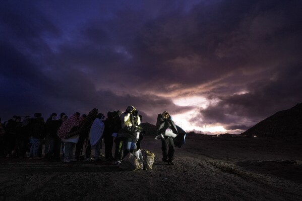 Asylum-seeking migrants wrap themselves in blankets to ward off the wind and rain as they line up in a makeshift, mountainous campsite to be processed after crossing the border with Mexico, Friday, Feb. 2, 2024, near Jacumba Hot Springs, Calif. (AP Photo/Gregory Bull)