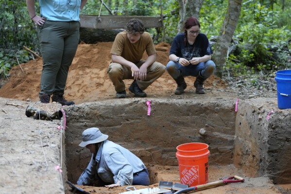 Gray Tarry, bottom left, an archeological field technician for the University of Louisiana at Lafayette, digs, while Josiah Hamilton, left, and Jamie Butts, right, high school students from Youth Conservation Corps, watch at an archeological site in Kisatchie National Forest, La., Wednesday, June 7, 2023. This summer, archaeologists have been gingerly digging up the ground at the site in Vernon Parish to unearth and preserve the evidence of prehistoric occupation. The site was found by surveyors in 2003, according to the U.S. Forest Service. Hurricanes Laura and Delta uprooted trees and exposed some of the artifacts. Further damage has been done by looters making unauthorized digs. Forest officials say the site shows evidence of generations of people living in the area going back 12,000 years. (AP Photo/Gerald Herbert)