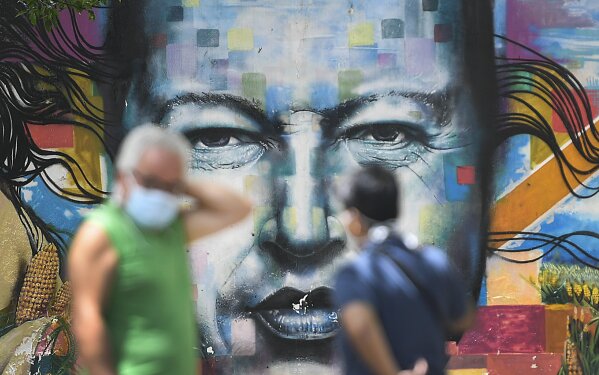 Pedestrians wearing masks to curb the spread of the new coronavirus chat in front of a mural of late President Hugo Chavez, in Caracas, Venezuela, Sunday, July 12, 2020. (AP Photo/Matias Delacroix)