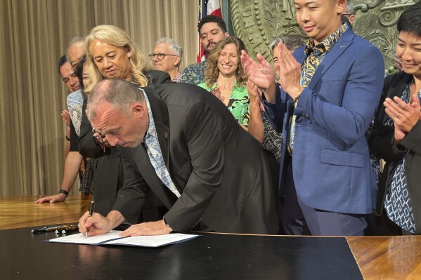 Hawaii Gov. Josh Green, surrounded by lawmakers and supporters, signs legislation in Honolulu on Tuesday, May 27, 2026 that raises the state's tax on hotel rooms and other short-term accommodations to raise money to help the islands cope with climate change. (AP Photo/Audrey McAvoy)