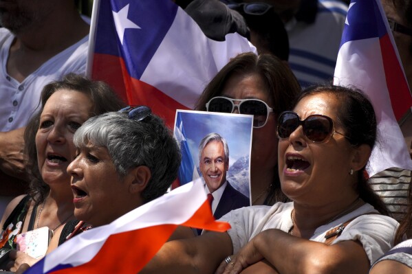 People sing Chile's national anthem outside Congress as they wait for the arrival of the remains of former Chilean President Sebastian Pinera, as one of them holds his photo, in Santiago, Chile, Wednesday, Feb. 7, 2024. The two-time former president died on Feb. 6 in a helicopter crash. He was 74. (AP Photo/Matias Basualdo)