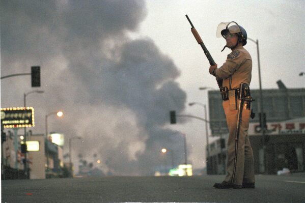 A California Highway Patrol officer stands guard at Ninth Street and Vermont Avenue in Los Angeles as smoke rises from a fire further down the street, April 30, 1992. (AP Photo/David Longstreath, file)
