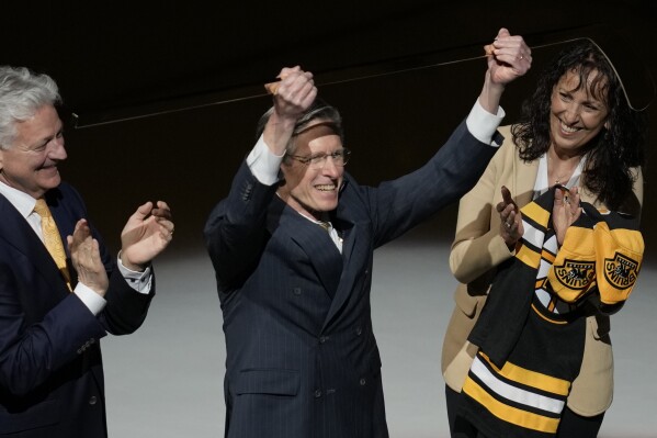 Boston Bruins television play-by-play announcer Jack Edwards, center, holds up a golden stick in honor of his announced retirement, before the Bruins' NHL hockey game against the Ottawa Senators, Tuesday, April 16, 2024, in Boston. (AP Photo/Charles Krupa)