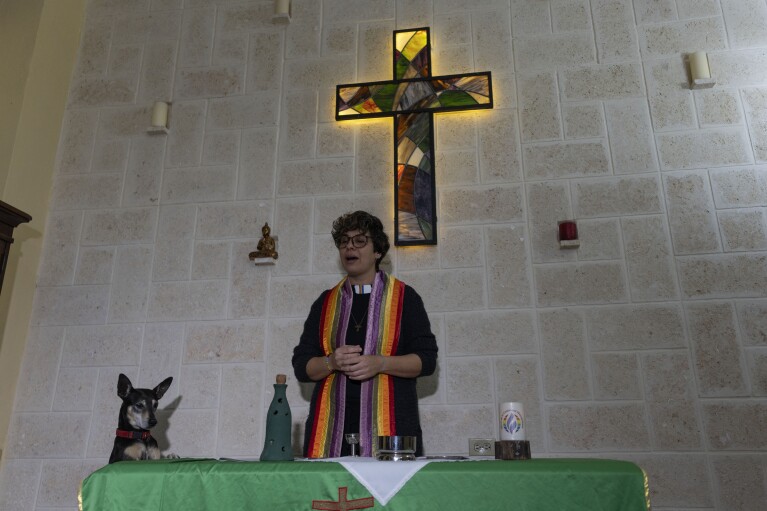 FILE - Rev. Elaine Saralegui, wearing a rainbow-colored clergy stole and her clerical collar, welcomes congregants to a service at the Metropolitan Community Church, an LGBTQ+ inclusive house of worship, as Ruth the dog stands with her front paws on the altar table, in Matanzas, Cuba, Feb. 2, 2024. In 2015, with support from the U.S.-based LGBTQ+ affirming Metropolitan Community Churches, they converted a house into their church, decked with wooden pews and a stained-glass cross that hangs above the altar. (AP Photo/Ramon Espinosa, File)