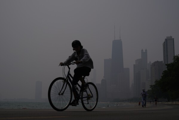 FILE - A person rides a bicycle along the shore of Lake Michigan as the downtown skyline is blanketed in haze from Canadian wildfires June 27, 2023, in Chicago. Forecasters say there won’t be large breaks for much of America anytime soon from eye-watering dangerous smoke from fire-struck Canada. (AP Photo/Kiichiro Sato, File)