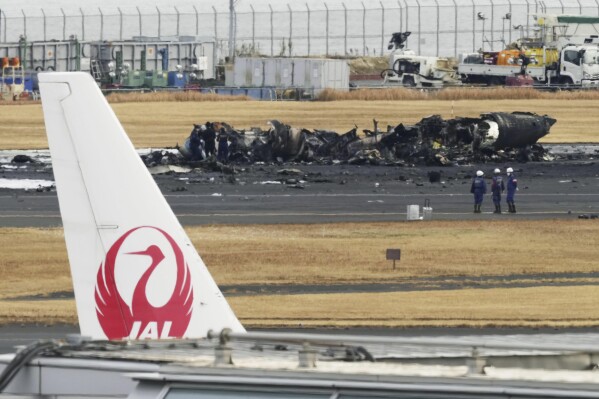 The burn-out Japanese coast guard aircraft is seen at rear behind the logo of Japan Airline at Haneda airport on Wednesday, Jan. 3, 2024, in Tokyo, Japan. (Kyodo News via AP)
