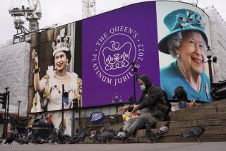 Un hombre alimenta a las palomas y a sus espaldas se ve una pantalla en Piccadilly Circus para celebrar el 70 aniversario del ascenso al trono de la reina Isabel de Gran Bretaña, en Londres, el domingo 6 de febrero de 2022. (Foto AP/Alberto Pezzali)