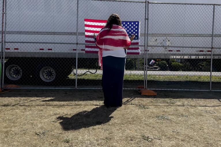 A woman prays as she faces a U.S. flag on the perimeter fence at the ReAwaken America Tour at Cornerstone Church in Batavia, N.Y., Saturday, Aug. 13, 2022. Anthea Butler, a scholar of American religion and politics at the University of Pennsylvania says that that the way Michael Flynn and ReAwaken join Christian nationalism to the idea of spiritual warfare is dangerous because it suggests there are “demonic” people in government, and Christians need to act to save the country. “If people are talking about spiritual warfare and are talking about taking up arms and stuff, then I think you should be very worried,” Butler says. (AP Photo/Carolyn Kaster)