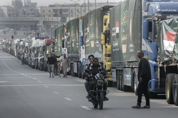 People walk past trucks of humanitarian aid at a parking point in Cairo, Egypt, as they wait to travel to cross the Rafah border crossing between Egypt and the Gaza Strip, Sunday, Jan. 26, 2025. (AP Photo/Amr Nabil)