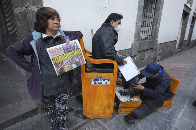 A newspaper vender holds a paper with the Spanish headline "Terrorists!" near her kiosk as a shoe shiner attends a client outside the Carondelet government palace, the presidential office and residence in Quito, Ecuador, Wednesday, Jan. 10, 2024. The headline refers to the previous day's attack on a TV station in the city of Guayaquil where masked gunmen burst into the studio, broadcast live. Authorities said the attackers were arrested and would be charged with terrorism. (AP Photo/Dolores Ochoa)