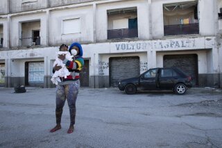 FILE -- In this photo taken on Monday, April 27, 2020, a woman wearing a sanitary mask to protect against COVID-19 holds her daughter as she walks past a building in Castel Volturno, near Naples, Southern Italy. They are known as “the invisibles,” the undocumented African migrants who, even before the coronavirus outbreak plunged Italy into crisis, barely scraped by as day laborers, prostitutes and seasonal farm hands. Pope Francis is calling for migrant farm workers to be treated with dignity, issuing an appeal as Italy weighs whether to legalize undocumented agricultural workers amid a shortage of seasonal farm labor due to the coronavirus emergency. (AP Photo/Alessandra Tarantino)