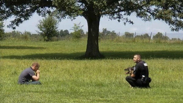 In this image from video provided by a family friend, Taylor Ware, left, sits in a field approached by a police officer and canine at a highway rest stop in Dale, Ind., on Aug. 25, 2019. Taylor's mother called 911 when he wouldn’t get back in their SUV during a manic episode caused by bipolar disorder. (Pauline Engel via AP)