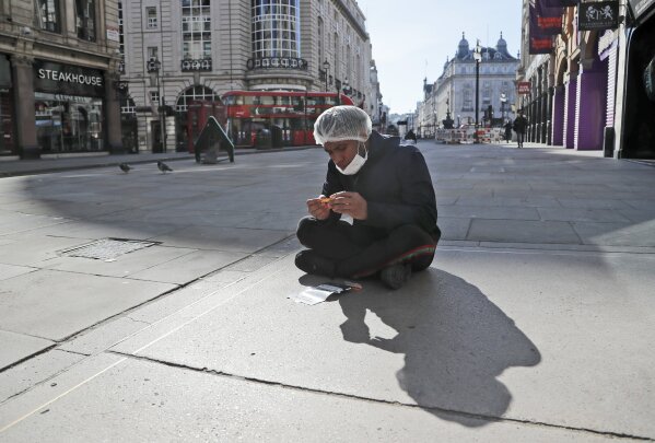 An employee of a nearby takeaway restaurant rolls a cigarette as he has a break sitting on the pavement near Piccadilliy Circus in central London, Tuesday, March 24, 2020.  Britain's Prime Minister Boris Johnson on Monday imposed its most draconian peacetime restrictions due to the spread of the coronavirus on businesses and social gatherings. For most people, the new coronavirus causes only mild or moderate symptoms. For some it can cause more severe illness.(AP Photo/Frank Augstein)
