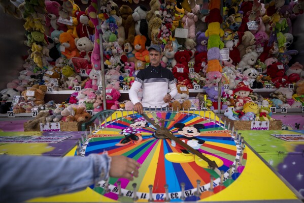 A man waits for customers to spin the wheel for winning plush toys at a fair in Hagioaica, Romania, Saturday, Sept. 16, 2023. For many families in poorer areas of the country, Romania's autumn fairs, like the Titu Fair, are one of the very few still affordable entertainment events of the year. (AP Photo/Andreea Alexandru)