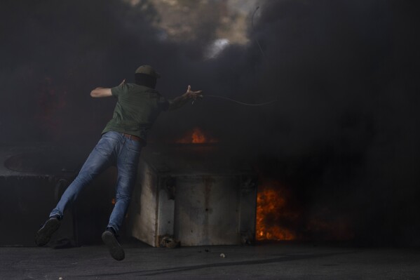 A Palestinian protester uses a slingshot behind burning tires during clashes with Israeli border police following a protest against Israeli airstrikes on Gaza, in the West Bank city of Ramallah, Wednesday, Oct. 18, 2023. (AP Photo/Nasser Nasser)