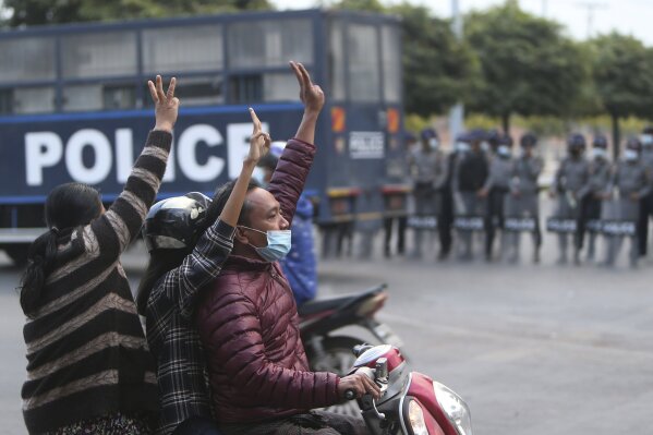 Protesters on a motorcycle flash the three-fingered salute as they drive past police in riot gear in Mandalay, Myanmar, on Feb. 6, 2021. Protests in Myanmar against the military coup that removed Aung San Suu Kyi’s government from power have grown in recent days despite official efforts to make organizing them difficult or even illegal. (AP Photo)