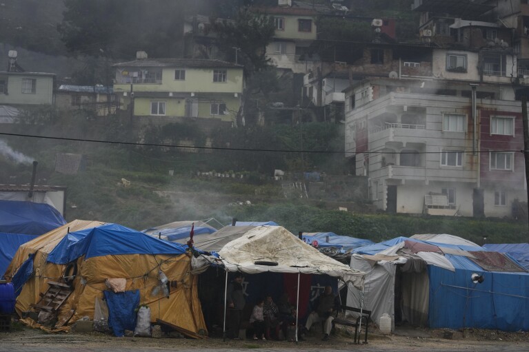 Neighbors sit and chat outside a tent at a makeshift campsite where families who are too afraid to return to their homes are sheltering in Antakya, southern Turkey, Friday, Jan. 12, 2024. Some families whose homes were listed as being “slightly damaged” in the powerful Feb. 6, 2023 earthquake have not been able to obtain temporary container homes. (AP Photo/Khalil Hamra)