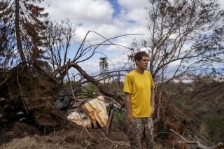 Kyle Ellison stands in front of a fallen tree on Wednesday, Sept. 27, 2023, in Kula, Hawaii. As high winds whipped burning trees and grass, Ellison and his landlord struggled with plummeting water pressure. Ellison had to wait for pots to slowly fill in the sink before running them to the fire; his landlord wielded a garden hose with little more than a trickle. (AP Photo/Mengshin Lin)