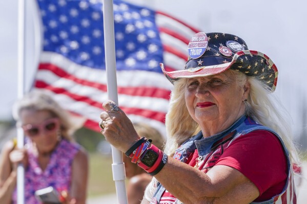 FILE - Supporters wave flags as they wait for the motorcade of former President Donald Trump to arrive at Palm Beach International Airport in West Palm Beach, Fla., March 25, 2023. Millions of Americans will attend parades, fireworks, barbecues and other Independence Day events on Tuesday, celebrating the courage and sacrifices of the nation’s 18th century patriots who fought for the nation’s independence from England and what they considered an unjust government. (AP Photo/Gerald Herbert, File)