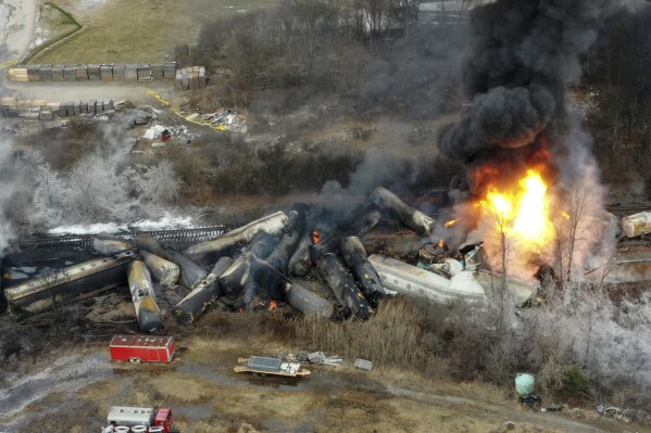 FILE - Portions of a Norfolk Southern freight train that derailed the night before burn in East Palestine, Ohio, Feb. 4, 2023. Norfolk Southern railroad announced on Tuesday, Dec. 5, plans to stop paying relocation aid to people displaced by the derailment right after the one-year anniversary of the crash. Railroad officials reiterated their long-term commitment to helping the town of East Palestine and the surrounding area recover. (AP Photo/Gene J. Puskar, File)