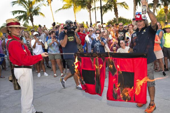 In this photo provided by the Florida Keys News Bureau, Jai Somers, left, holds burning hurricane flags after they were doused with rum and set on fire by Paul Menta, right, to mark the end of the 2022 Atlantic hurricane season Wednesday, Nov. 30, 2022, in Key West, Fla. According to the National Hurricane Center, the 2022 season spawned 14 named tropical cyclones including eight hurricanes with two classified as major hurricanes. The event, which attracted several hundred Florida Keys residents, was staged by Key West's ceremonial Conch Republic "government." (Andy Newman/Florida Keys News Bureau via AP)