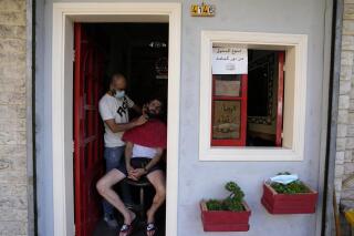 A Lebanese barber shaves the beard of a customer at the door of his shop during a power outage in a southern suburb of Beirut, Lebanon, Wednesday, Aug. 11, 2021. Lebanon, which is mired in multiple crises including a devastating economic crisis, has faced months of severe fuel shortages that have prompted long lines at gas stations and plunged the small country, dependent on private generators for power, into long hours of darkness. Sign on door says, "Forbidden to enter without mask." (AP Photo/Hassan Ammar)