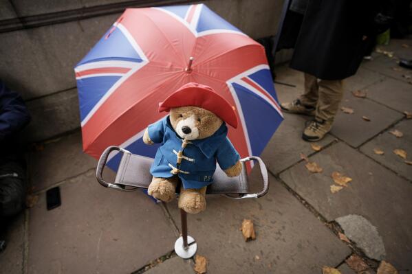 FILE - A Paddington bear stuffed toy is placed on a chair next to a Union flag umbrella as people wait opposite the Palace of Westminster to be first in line bidding farewell to Queen Elizabeth II in London on Sept. 14, 2022. More than 1,000 Paddington bears and other teddies left in tribute to the late queen in London and Windsor will be donated to a children's charity, Buckingham Palace said Saturday, Oct. 15. (AP Photo/Andreea Alexandru, File)