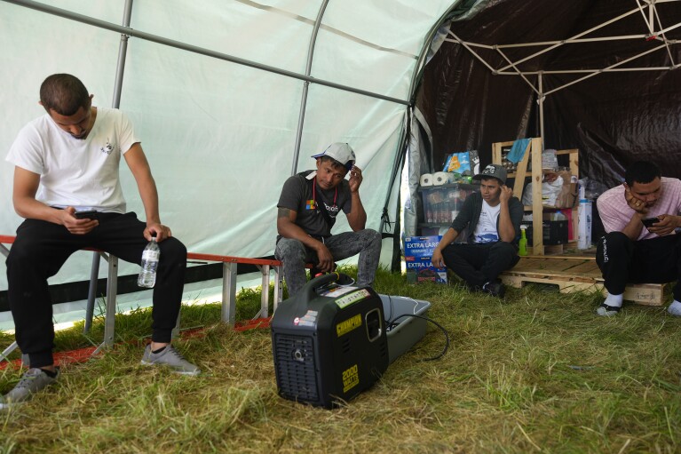 Men sit in the shade of a community tent near a charging station at an encampment of asylum-seekers mostly from Venezuela, Congo and Angola next to an unused motel owned by the county, Wednesday, June 5, 2024, in Kent, Washington. The group of about 240 asylum-seekers is asking to use the motel as temporary housing while they look for jobs and longer-term accommodations. (AP Photo/Lindsey Wasson)