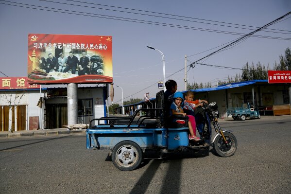 
              FILE - In this Sept. 20, 2018, file photo, an Uighur woman shuttles school children on an electric scooter as they ride past a propaganda poster showing China's President Xi Jinping joining hands with a group of Uighur elders in Hotan, in western China's Xinjiang region. Under Chinese President Xi Jinping, the Uighur homeland has been blanketed with stifling surveillance, from armed checkpoints on street corners to facial-recognition-equipped CCTV cameras steadily surveying passers-by. Now, Uighurs say, they must live under the watchful eye of the ruling Communist Party even inside their own homes. (AP Photo/Andy Wong, File)
            