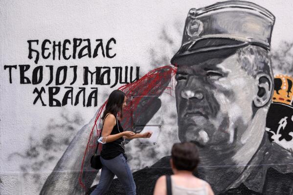 People walk by a mural of former Bosnian Serb military chief Ratko Mladic vandalized with red paint in Belgrade, Serbia, Saturday, July 24, 2021. The Bosnian Serb wartime political leader Radovan Karadzic and his military commander Ratko Mladic were both convicted of genocide in Srebrenica by a special U.N. war crimes tribunal in The Hague. Valentin Inzko, the outgoing head of Bosnia's Office of the High Representative, or OHR, imposed changes Friday to the country's criminal code, introducing prison sentences of up to five years for genocide denial and for the glorification of war criminals. (AP Photo/Darko Vojinovic)