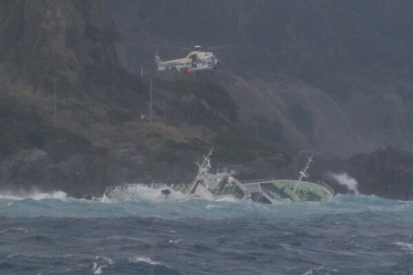 In this photo provided by Japan Coast Guard, a coast guard helicopter hovers over a fishing boat tilting almost to the side, for their rescue, off Kozushima, southwest of Tokyo, Monday, March 4, 2024. A tuna fishing boat went aground near one of Izu islands southwest of Tokyo after an engine trouble. ( Japan Coast Guard via AP)