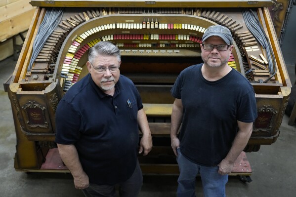 Carlton Smith, left, and Justin Nimmo pose in from of the console of the Barton Opus 234 theater organ that is undergoing restoration at Carlton Smith Pipe Organ Restorations in Indianapolis, Tuesday, July 2, 2024. The massive pipe organ built nearly a century ago for an extravagant movie house in Detroit is expected to be the centerpiece of a performing arts center at the Rochester Institute of Technology in Rochester, N.Y. (AP Photo/Michael Conroy)