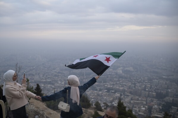 A young woman poses for a photo waving a Syrian "revolutionary" flag at a lookout area on the Mount Qasioun in Damascus, Syria, Saturday, Dec. 21, 2024. (AP Photo/Leo Correa)