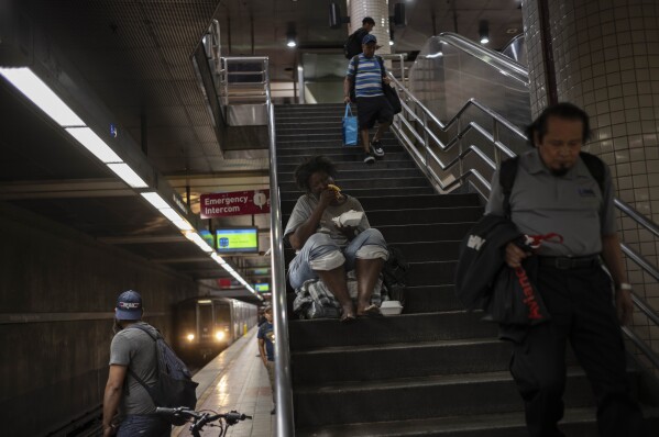 Commuters walk down the steps to board a Metro train as a homeless woman eats her food in Los Angeles, Tuesday, Sept. 19, 2023. (AP Photo/Jae C. Hong)