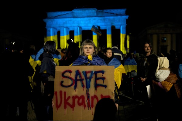 A protestor holds a placard at the Brandenburg Gate, illuminated in Ukrainian colors, during a demonstration against Russia's war on Ukraine as they mark the third anniversary of the full-scale invasion in Berlin, Monday, Feb. 24, 2025. (AP Photo/Ebrahim Noroozi)