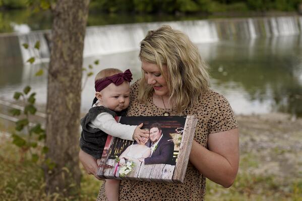 Aubrea Baker displays her wedding photo as she and her 7-month-old daughter Haylen visit one of her late husband's favorite fishing spots Saturday, Oct. 2, 2021, in Burlington, Kan. Her husband, Danny Baker, was among the 700,000 U.S. victims of COVID-19, dying on Sept. 14 after testing positive in July. (AP Photo/Charlie Riedel)