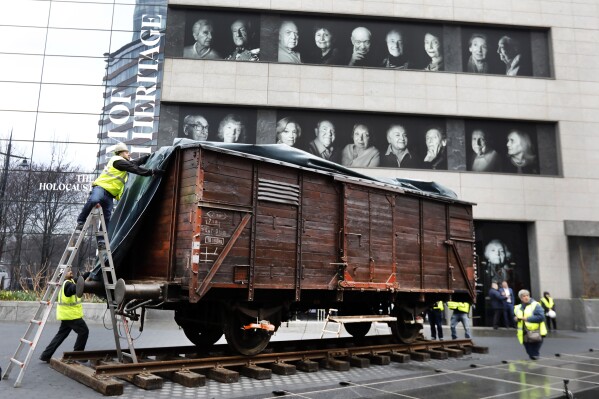 FILE - Portraits of Holocaust survivors are displayed at the Museum of Jewish Heritage as a vintage German train car, like those used to transport people to Auschwitz and other death camps, is uncovered on tracks outside the museum, in New York, Sunday, March 31, 2019. A privately funded effort in New York City will offer a Holocaust education field trips to all eighth graders in public schools. The partnership between the Museum of Jewish Heritage and Gray Foundation was announced Thursday by New York City Councilmember Julie Menin. (AP Photo/Richard Drew, File)