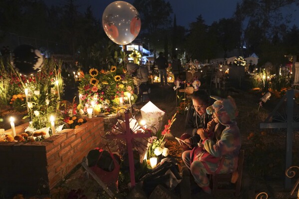 People sit around a child's tomb in the San Gregorio Atlapulco cemetery during Day of the Dead festivities on the outskirts of Mexico City, early Wednesday, Nov. 1, 2023. In a tradition that coincides with All Saints Day on Nov. 1 and All Souls Day on Nov. 2, families decorate graves with flowers and candles and spend the night in the cemetery, eating and drinking as they keep company with their dearly departed. (AP Photo/Marco Ugarte)