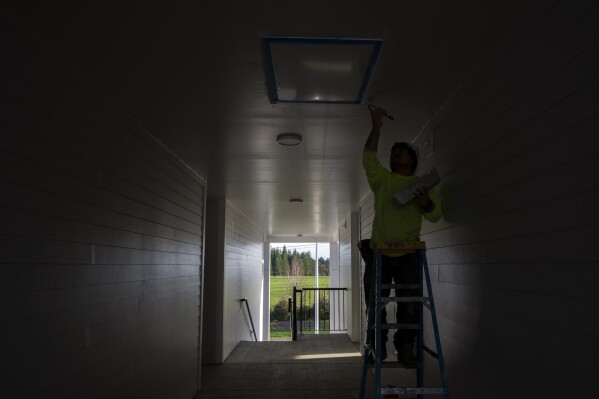 With the so-called urban growth boundary in the background, a worker paints the ceiling of the third-floor exterior corridor of a new construction complex on Thursday, Feb. 22, 2024, in the southwest Portland, Ore., suburb of Beaverton. The boundary was established by a 1973 law that placed boundaries around cities to prevent urban sprawl and preserve nature and farmland. (AP Photo/Jenny Kane)