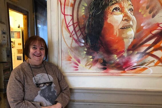 In this undated photo provided by Christian Guemy, Viviane Bouculat poses by a portrait of her by street artist Christian Guemy, known as C215, late in 2019, and painted directly on the wall of restaurant l'Annexe, in the Paris suburb of Ivry-sur-Seine. For three decades, Viviane Bouculat was the owner, cook and beating heart of l’Annexe. Anyone was welcome, but over the years it became a haven for local artists, actors and musicians. Bouculat died March 31, 2020 of coronavirus. She was 65. (Christian Guemy via AP)